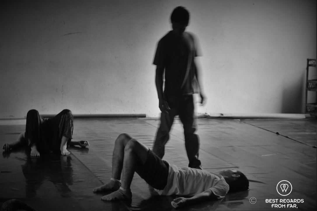2 pupils laying on the floor at the gymnastic hall being taught acrobatics by a teacher. standing in between them