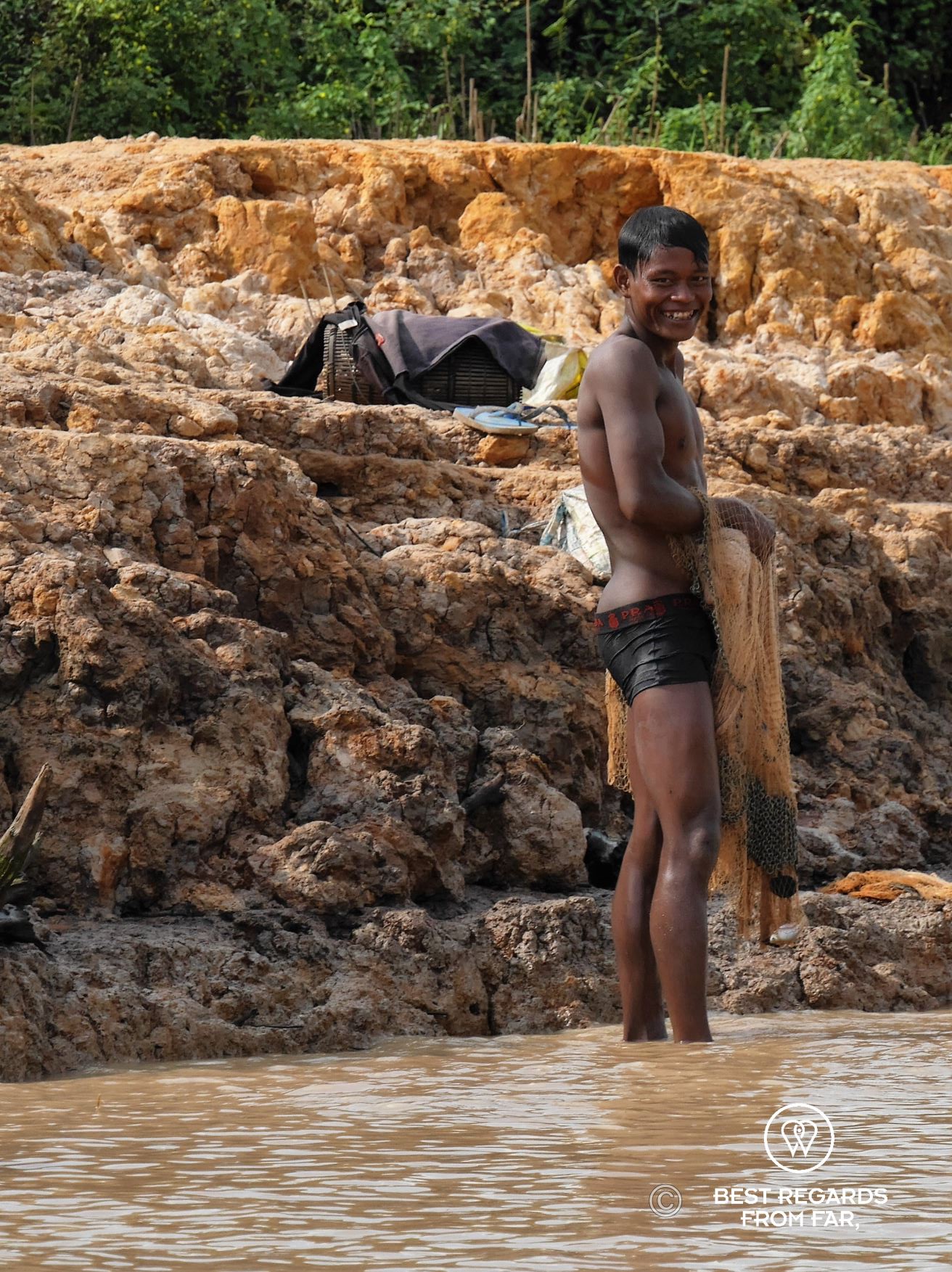 Smiley fisherman in swimming pants holding a net while standing in murky water.