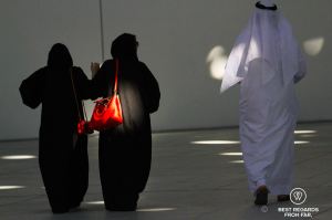 Two women walking wearing black burkas with red handbags and a man walking with a white thawb.