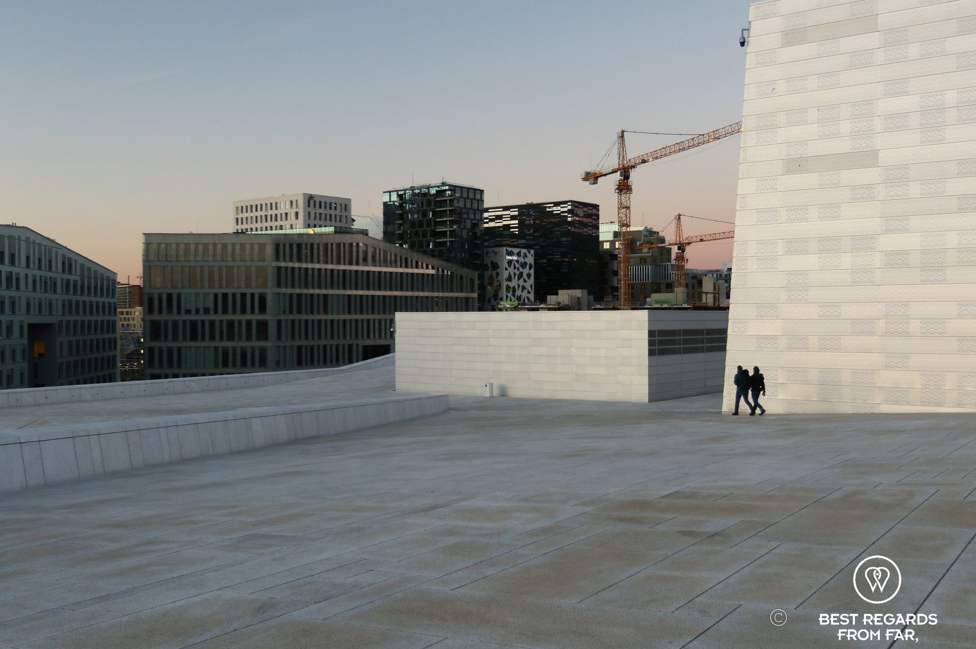 Two people walking on the white marble rooftop of the Oslo Opera House with modern buildings at the background.