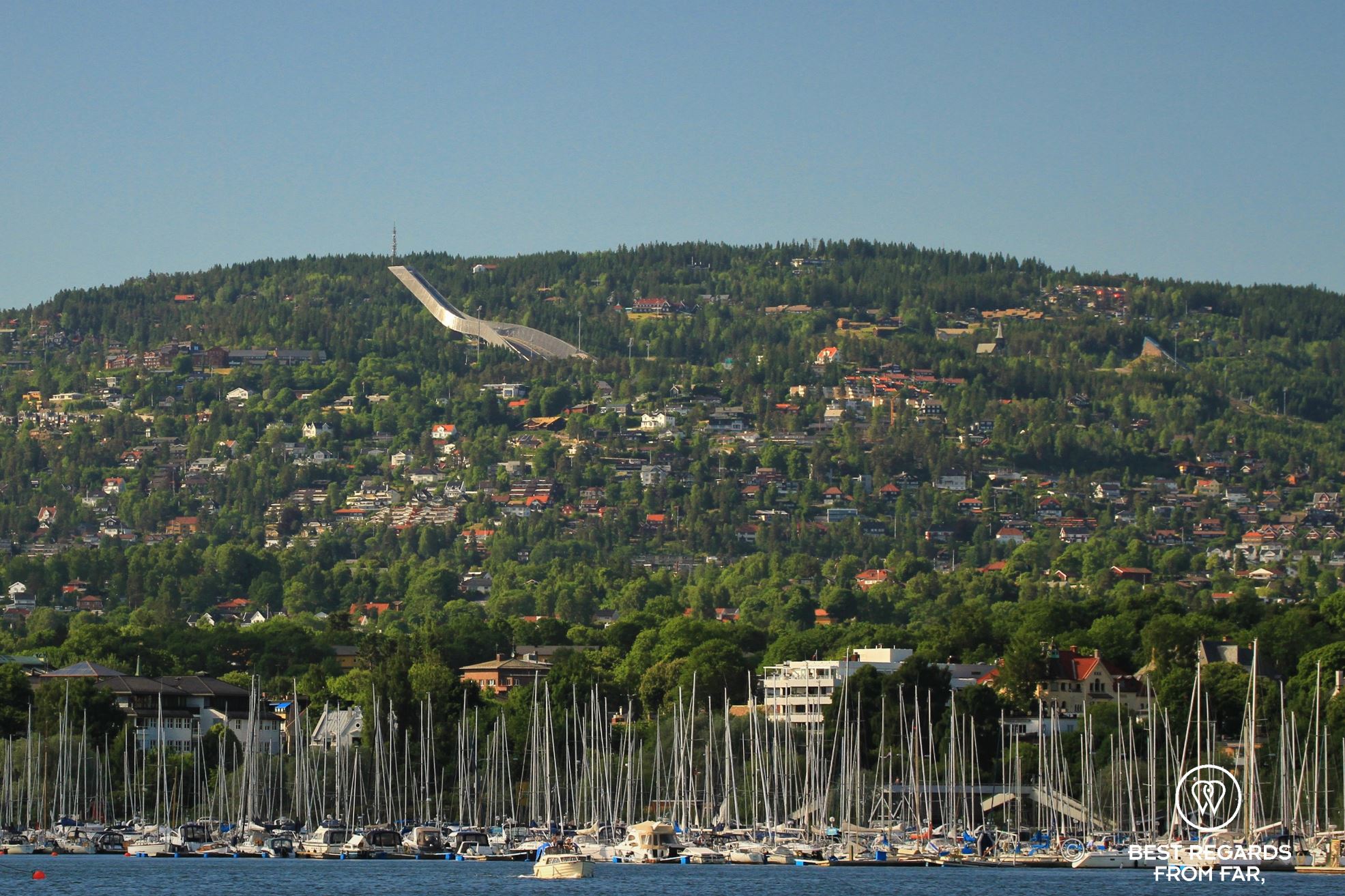 Blue water with a harbour full of sail boats, green mountain with a large ski jump, Oslo, Norway.