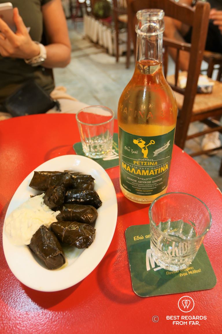 Red table with a bottle of Retsina, two glasses and a white plate of dolmades.