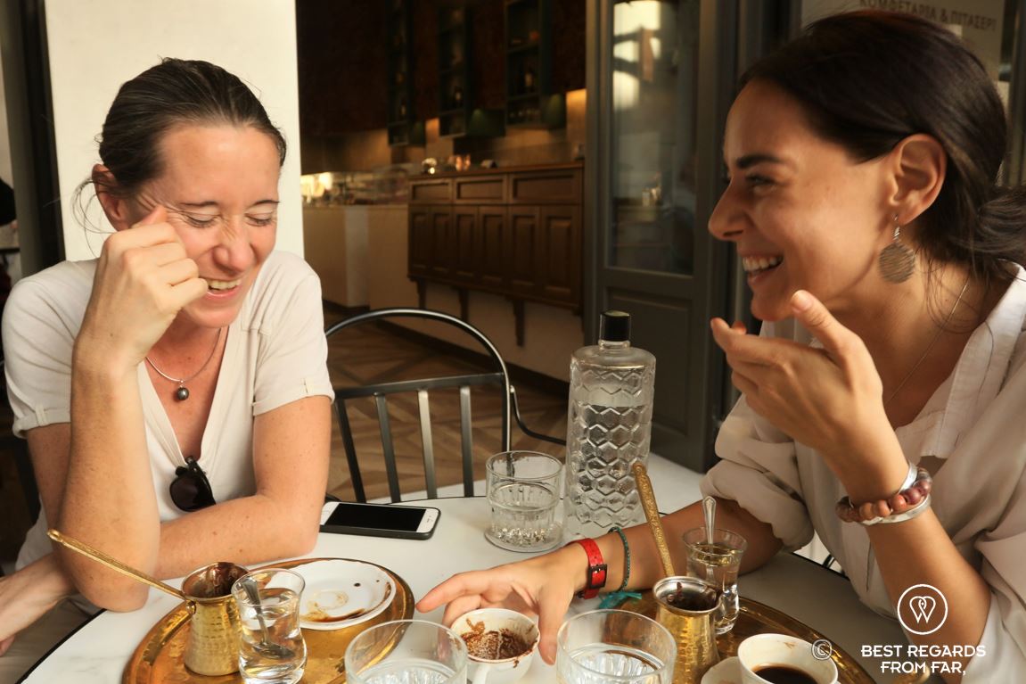 Two women laughing while reading coffee on a terrace in Greece.