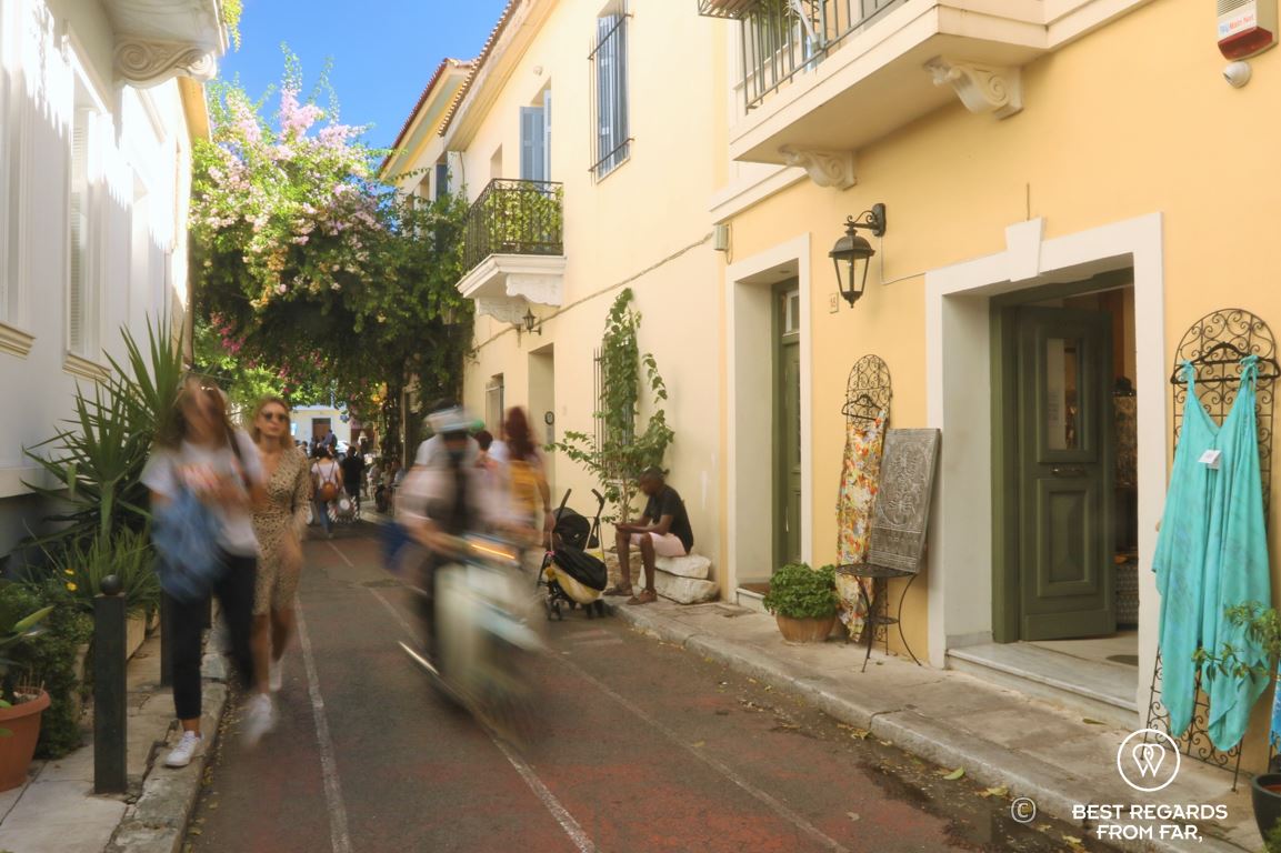 Two women walking a street, a scooter driving by and a blue dress on display in a street in Plaka, Athens, Greece.