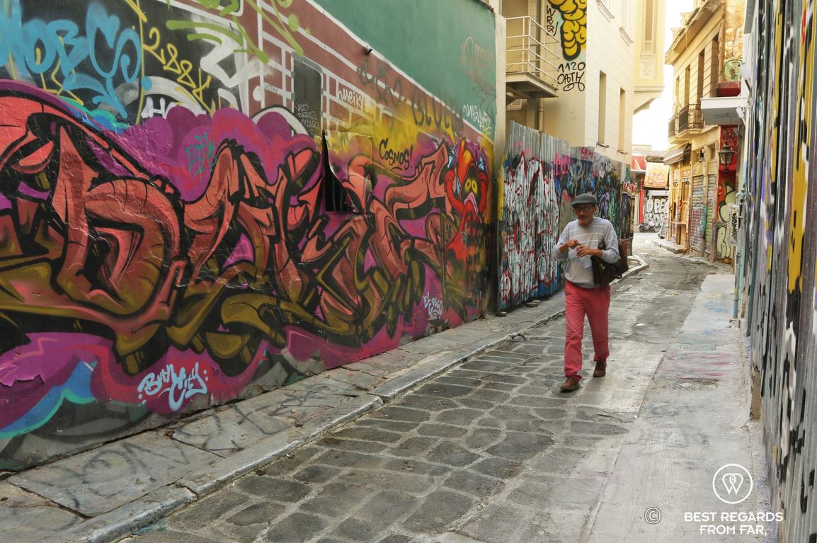 A man with red pants, grey jumber and green cap walking through a street covered in graffiti, Athens, Greece.