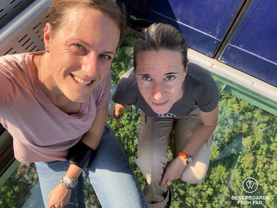 Two women in cacle car with see-through floor hovering over tree tops.