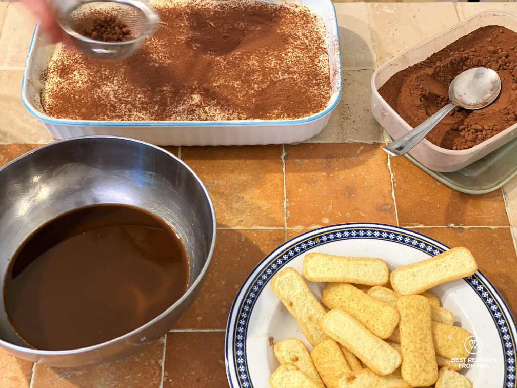 Recipe of tiramisu: adding cacao powder before presenting the tiramisu, with ingredients around (coffee, lady fingers, cacao).