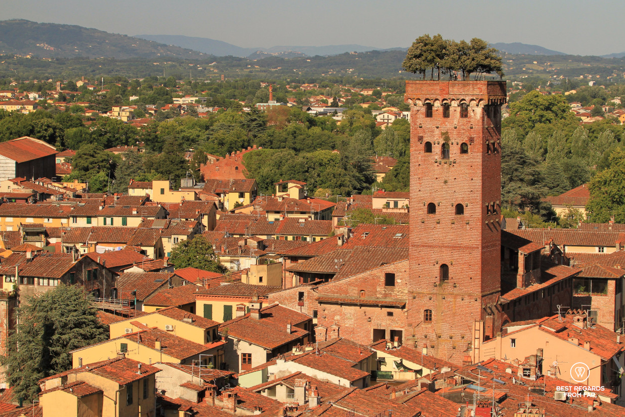 View on rooftops and a brick tower with trees on top of it and hills in the background