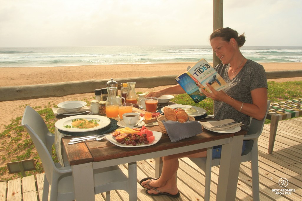 Woman on a wooden deck by the ocean reading and having breakfast