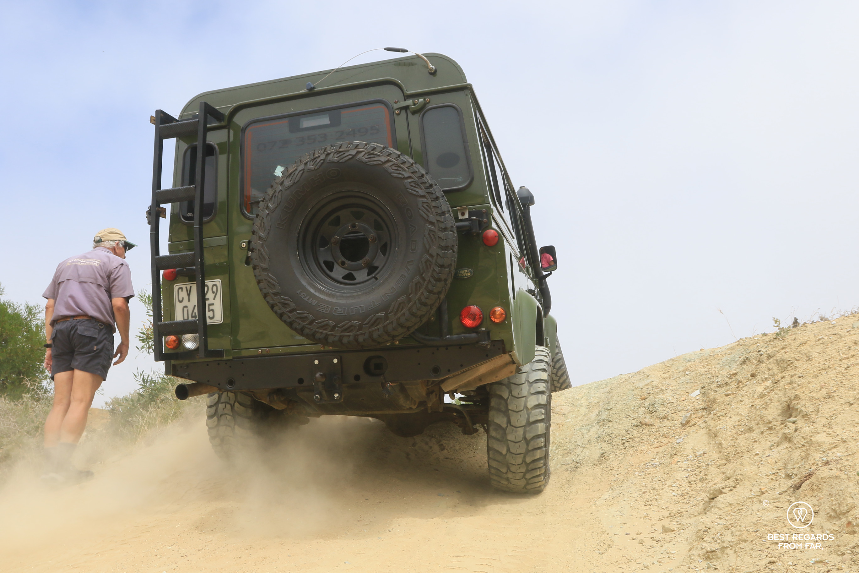 The rear of a green Land Rover Defender driving up a steep slope with the instructor walking next to it