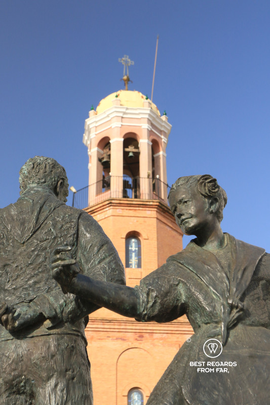 Bronze sculpture of a man and woman in front of the church of Competa with a bright blue sky.