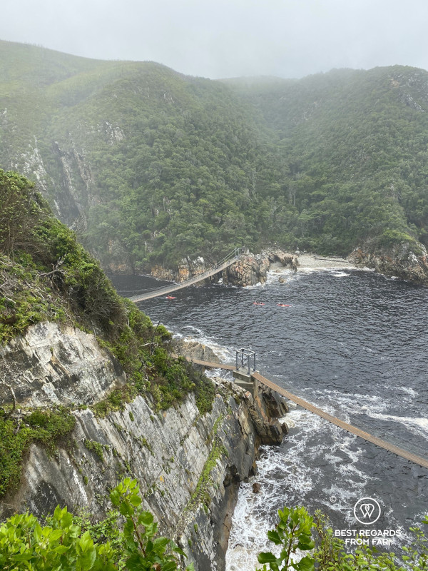 Two suspension bridges above the Indian Ocean along rocky mountains with green trees.
