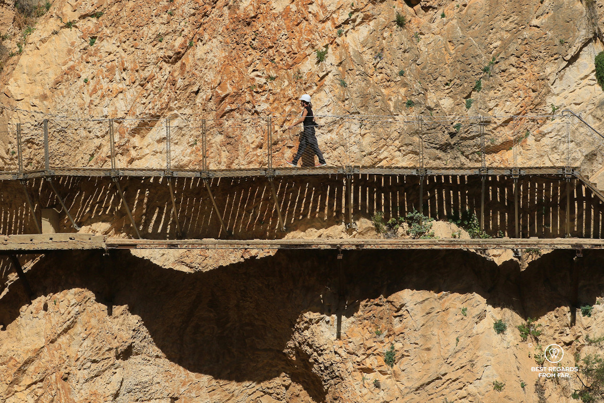 Woman wearing a white helmet walking a wooden boardwalk of Caminito del Rey bolted into a cliff.
