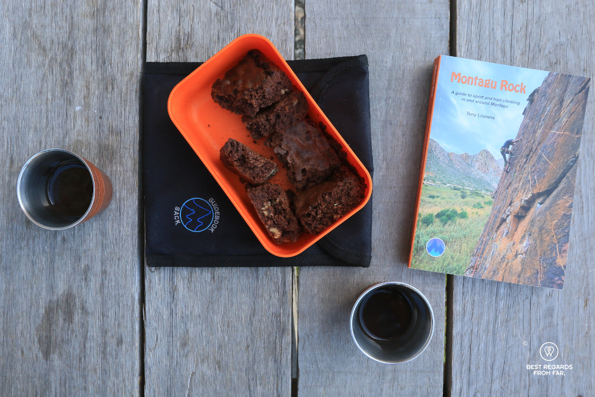 Two coffee cups with coffee, a brownie and the Montagu Rock book photographed from above on a wooden platform.