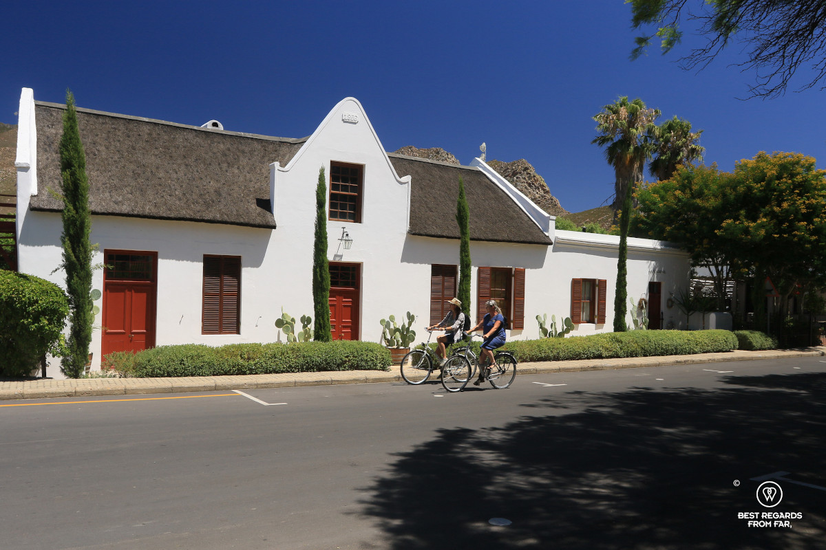 Two women biking by a white-washed historic house with a thatched roof in Montagu.