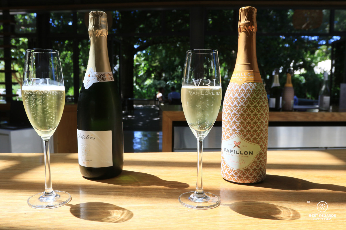 Two glasses of MCC with their bottles presented on a bar in the sun.