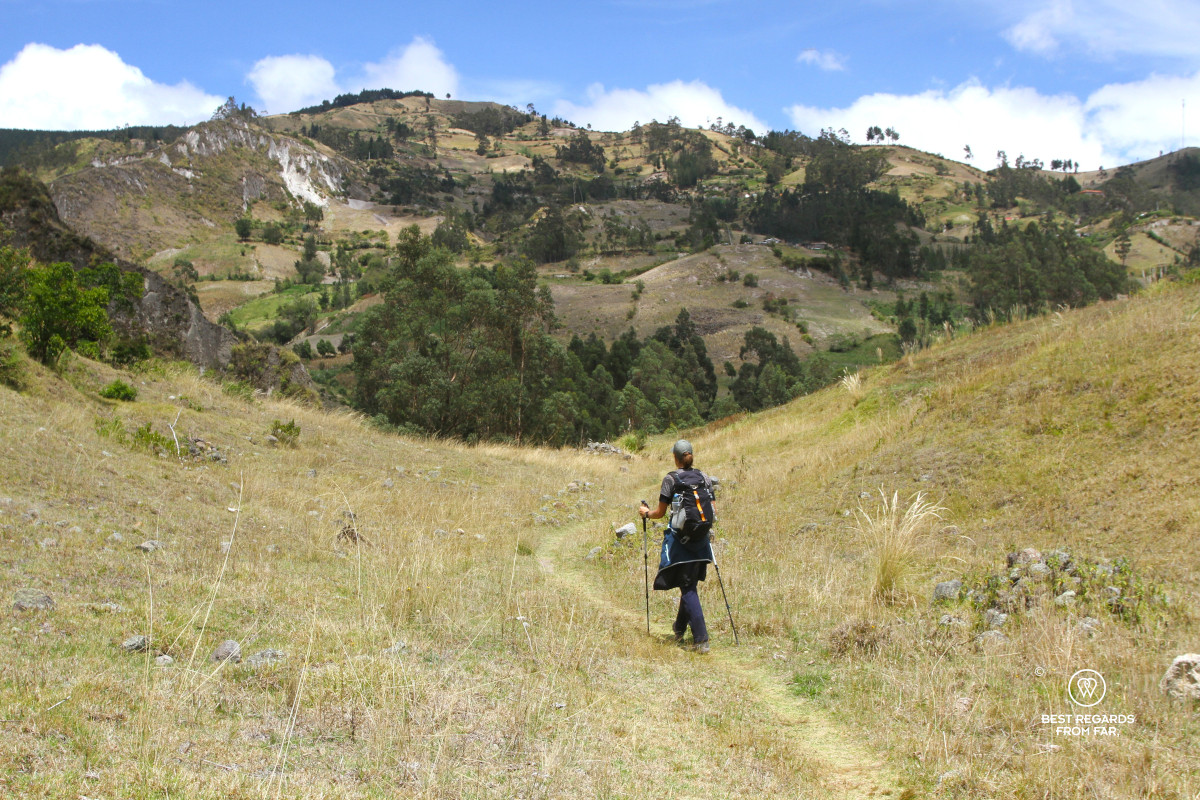Woman hiking on a grassy trail through a mountainous landscape.