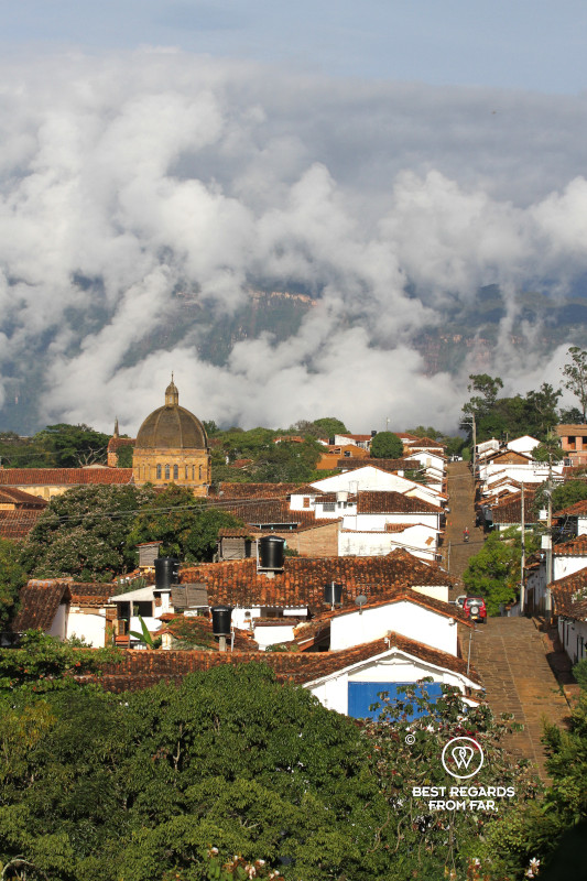 View on a cobblestoned street of Barichara with the church and rooftops and clouds in the background.