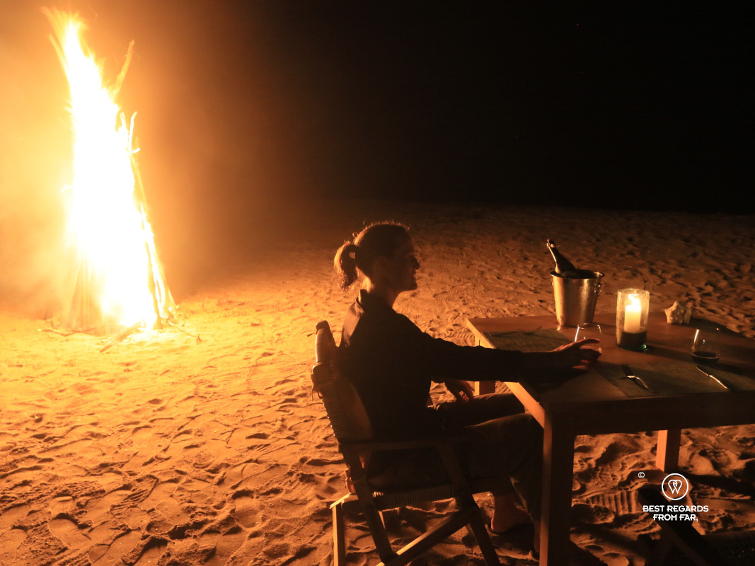 Woman at a table on the beach for dinner lit by a bonfire.