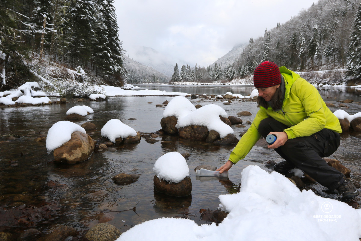 Woman in a green jacket filtering water with an ÖKO filter in the Jacques Cartier River with fir trees covered in snow.