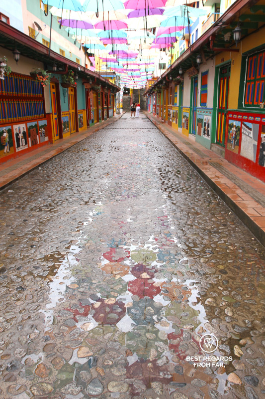 A colorful street in Guatape covered by colorful umbrellas that reflect in poddles.