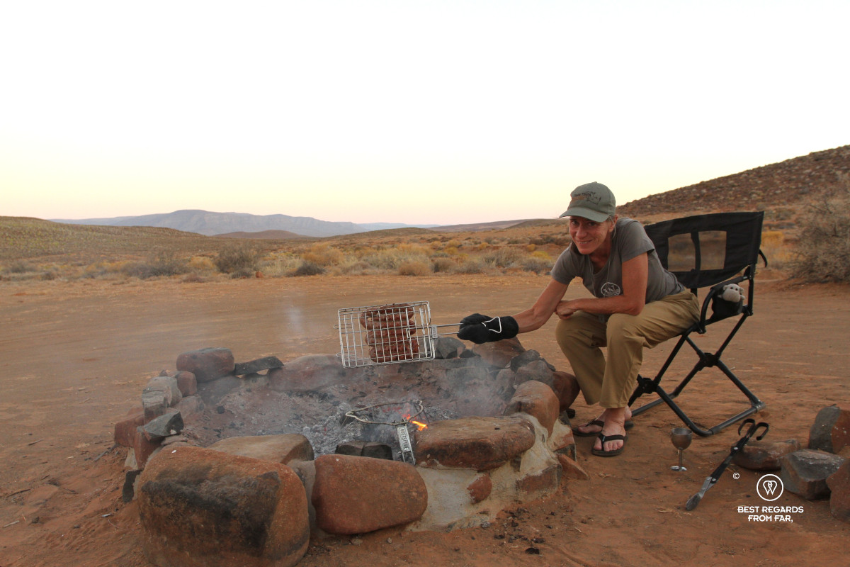 Woman holding grilled sausage above a fire pit in a barren landscape.