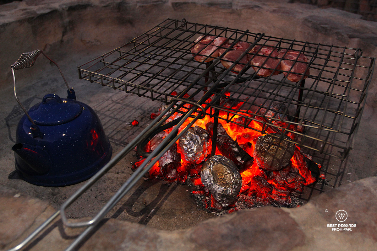 Lamb chops on the grill and potatoes amongst glowing coals with a tea pot to warm water next to it.
