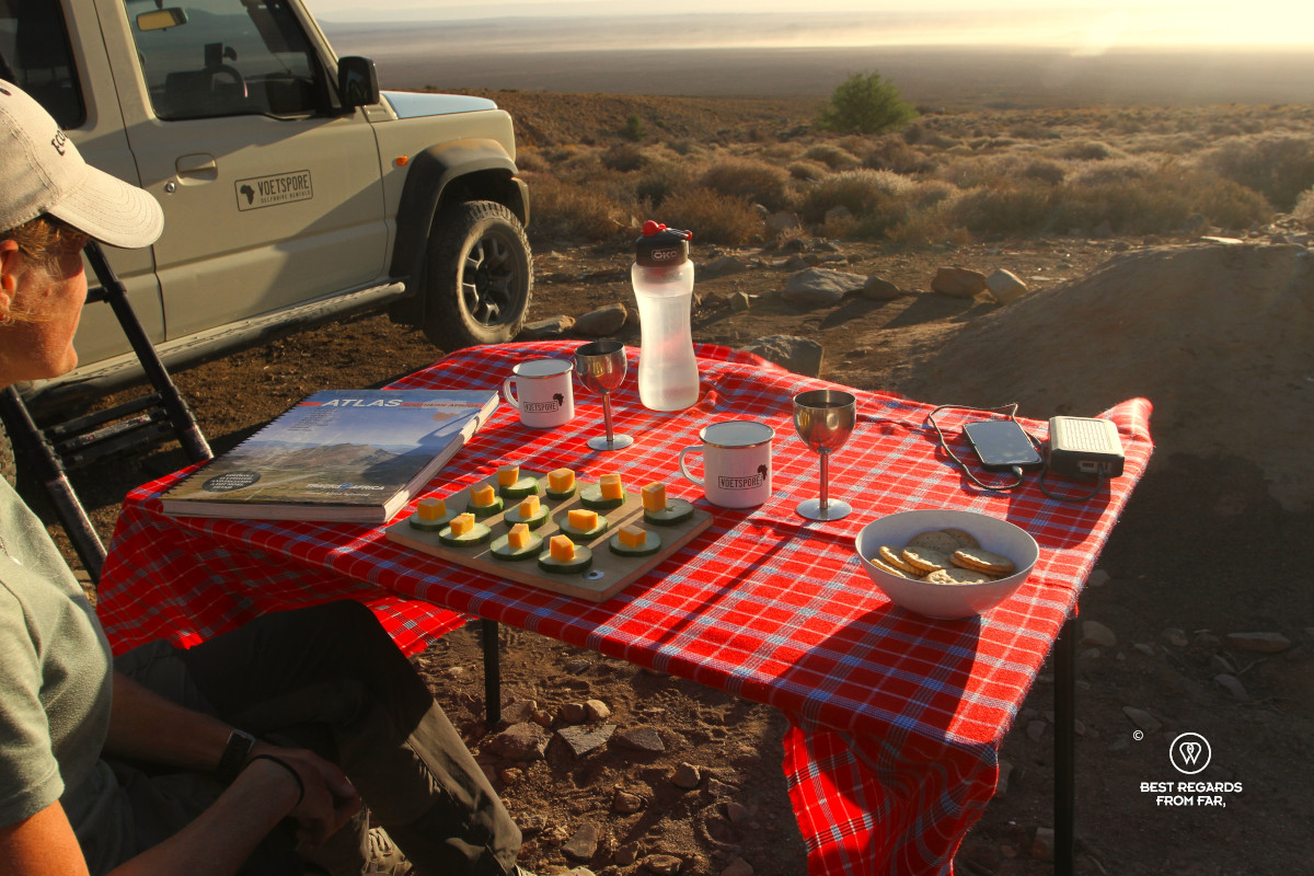 Woman taking in sunset next to her Suzuki Jimny with a set table and Tracks 4 Africa atlas.