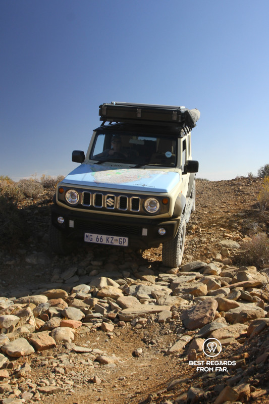 Geared up Suzuki Jimny approaching a dry river bed of stones.