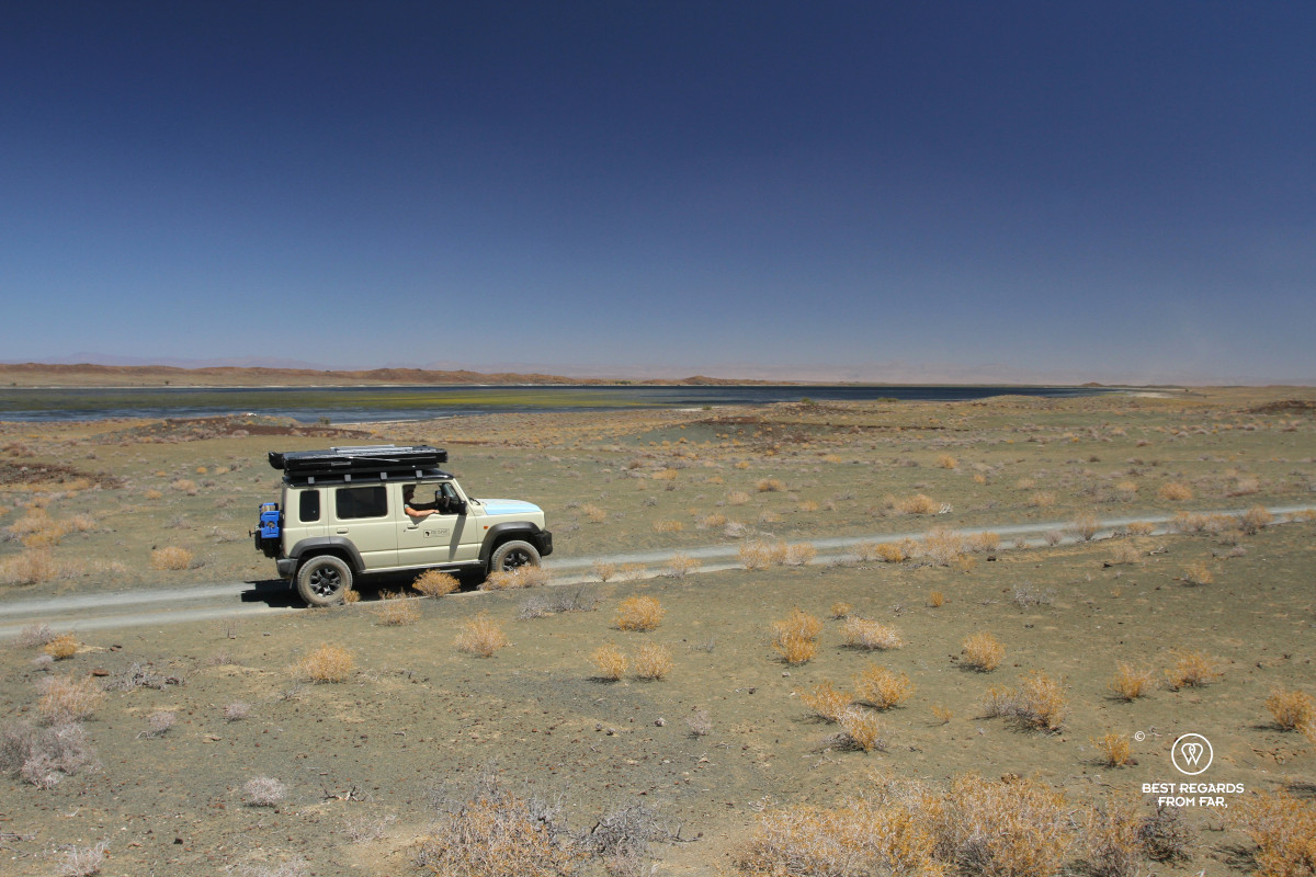 Beige Suzuki Jimny driving on a gravel road in front of a lake.