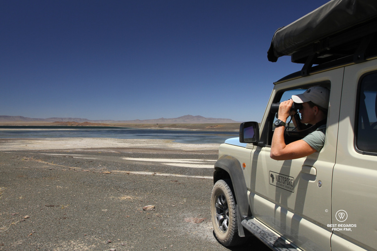 Woman taking in the view through binoculars from the passenger's seat of a beige Suzuki Jimny branded by Voetspore.