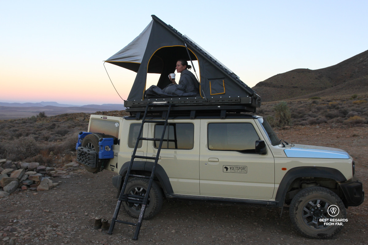 Woman in rooftop tent on a beige Suzuki Jimny having coffee at sunrise.