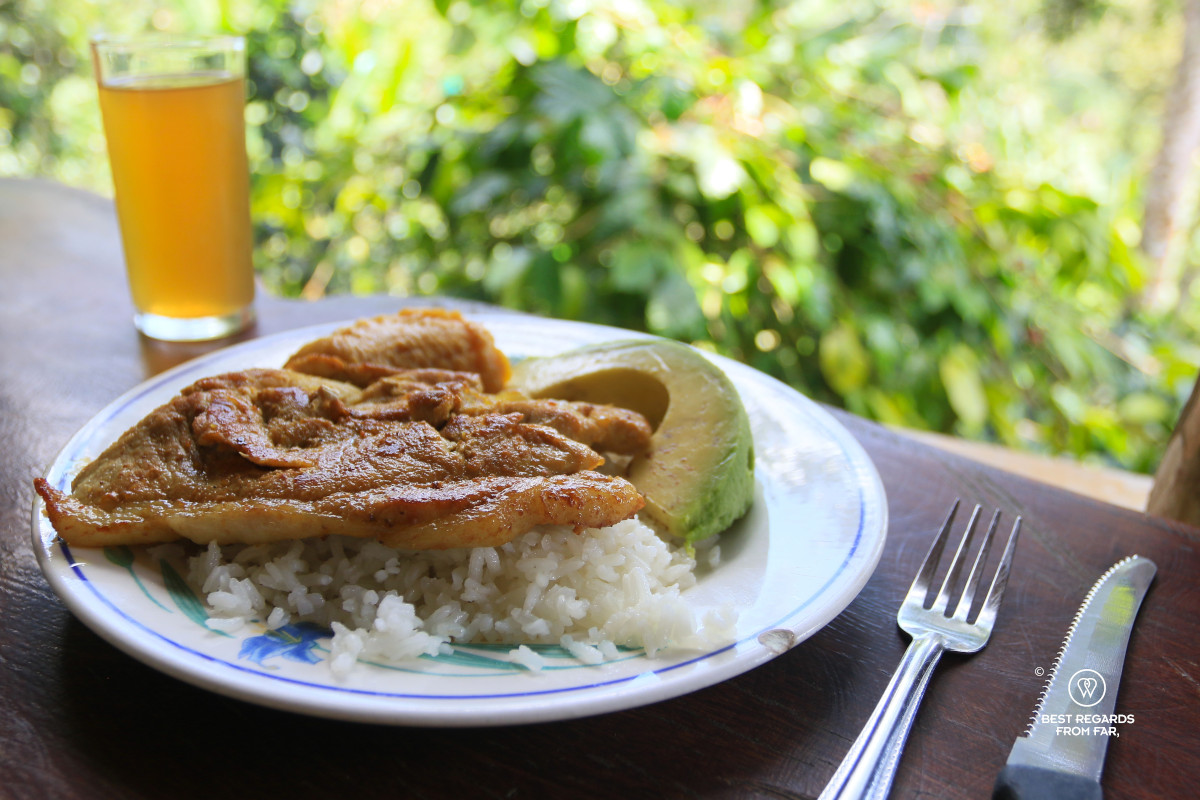 Plate on a wooden table with rice, meat, plantain and avocado and a glass of fruit juice.