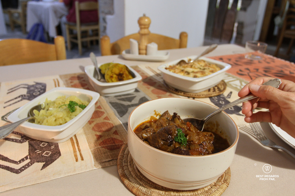 Table with different dishes for dinner and a meat dish in the foreground with someone scooping up a stew with a spoon.