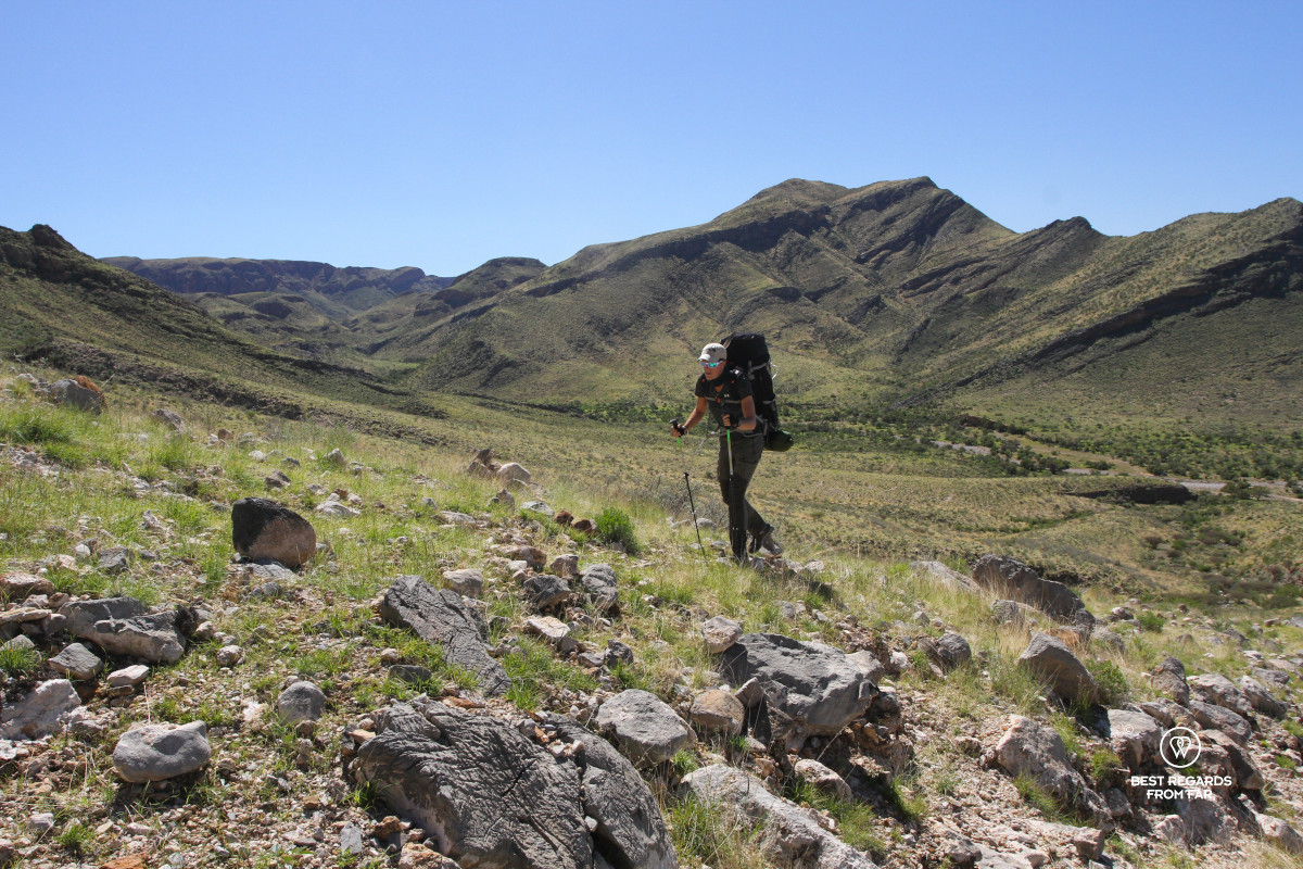 Woman with large backpack hiking through a green landscape with mountains in the background and blue skies above.