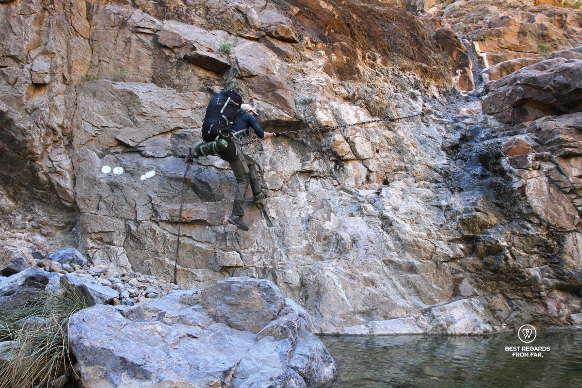 Author Marcella van Alphen carrying a large backpack while descending a canyon wall with the help of a chain.