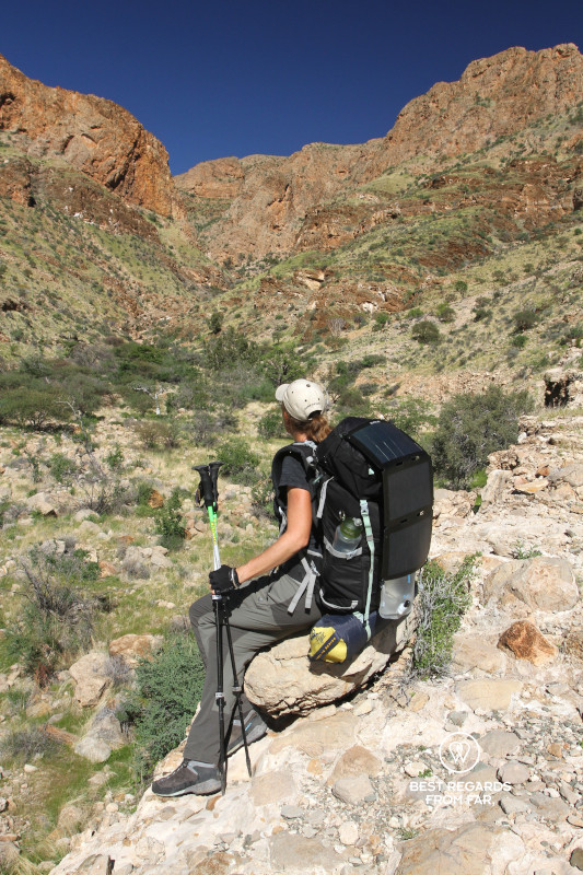 Author Marcella van Alphen in hiking outfit and with backpack resting on a rock overlooking a green valley and red mountains.