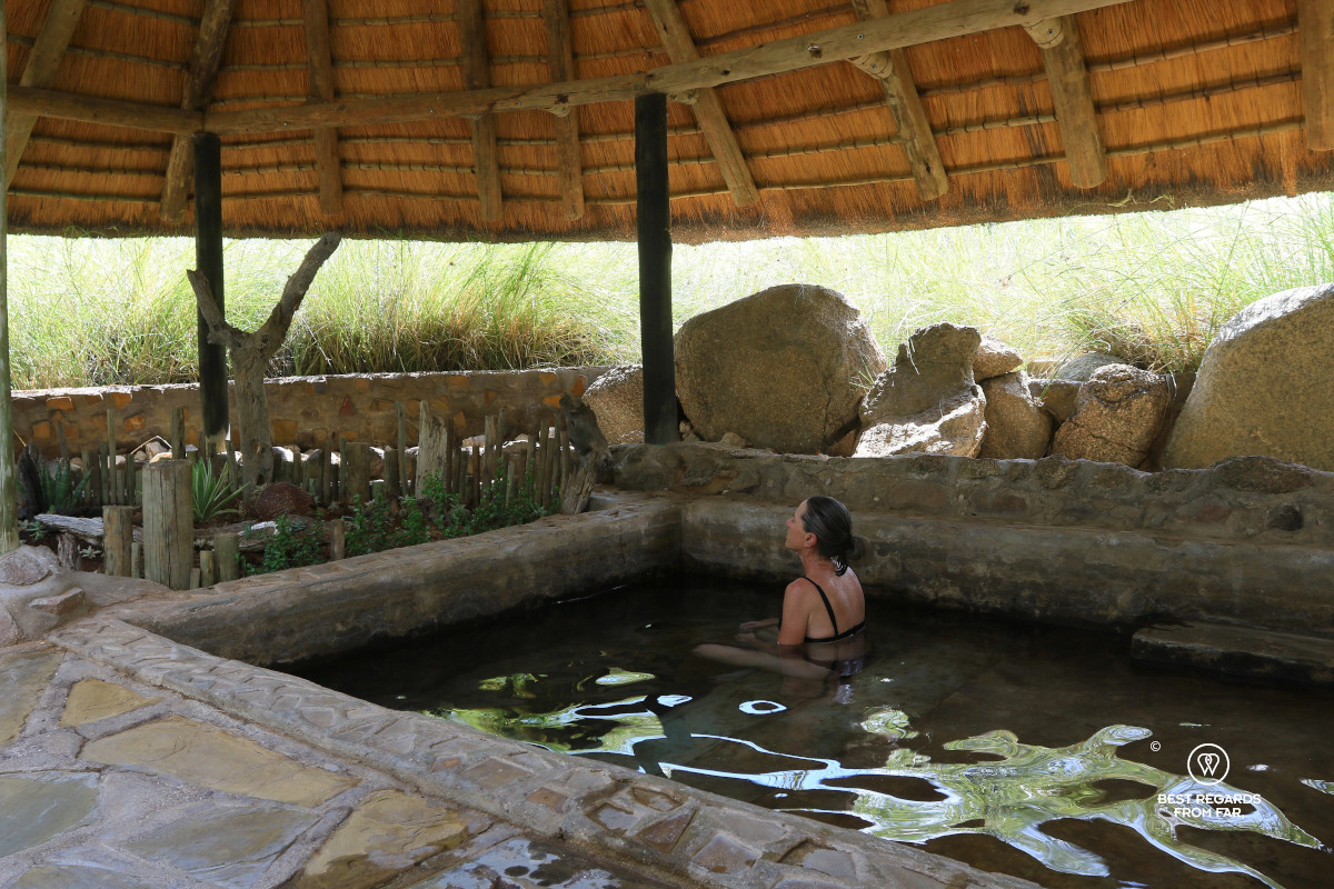 Author Claire Lessiau meditating in natural warm water spring with a thatched roof above it.