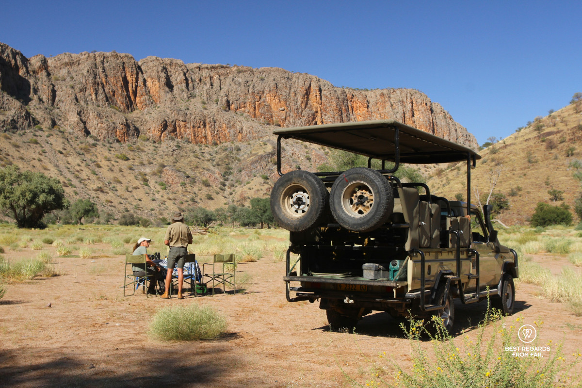 Toyota Land Cruiser parked in dry riverbed with man and woman by a set table and foldable chairs surrounded by red cliffs.