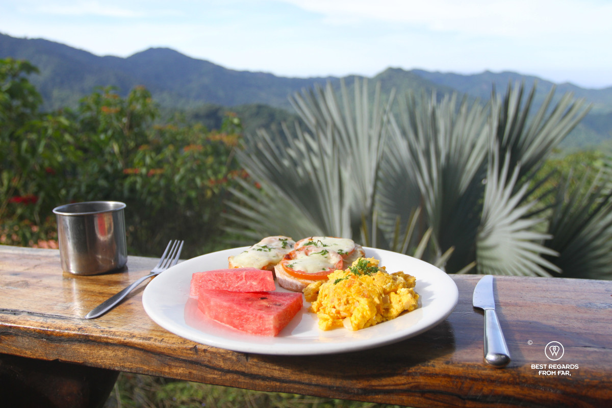White breakfast plate with eggs, watermelon and toast on wooden table overlooking green mountains.