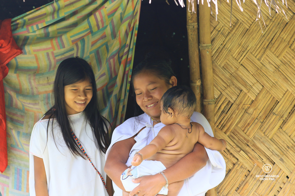 Wiwa mother holding her baby and a girl in front of their home made of natural materials.