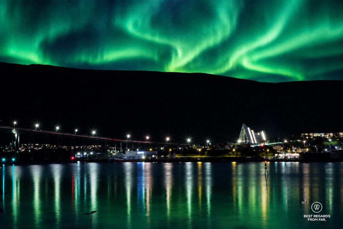 View on the Arctic Cathedral in Tromso with a green northern light dispaly behind the mountains at night.