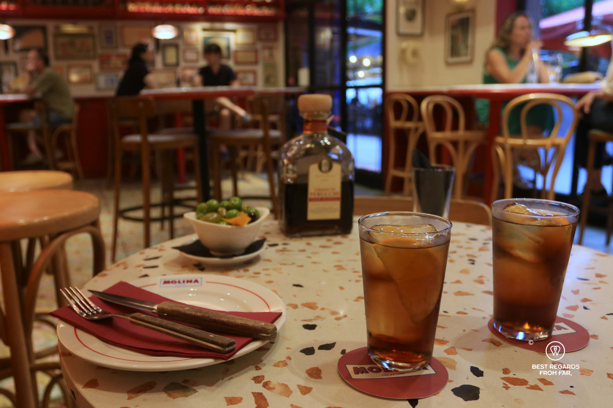 Two glasses of vermouth on a table with a bottle and olives.